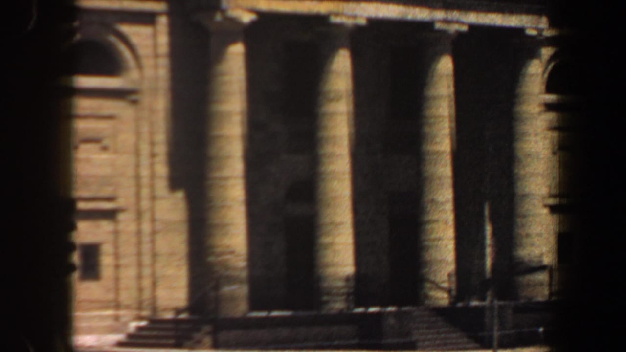 Columned building entrance with tan masonry, arched side detail, steps, and heavy shadow.
