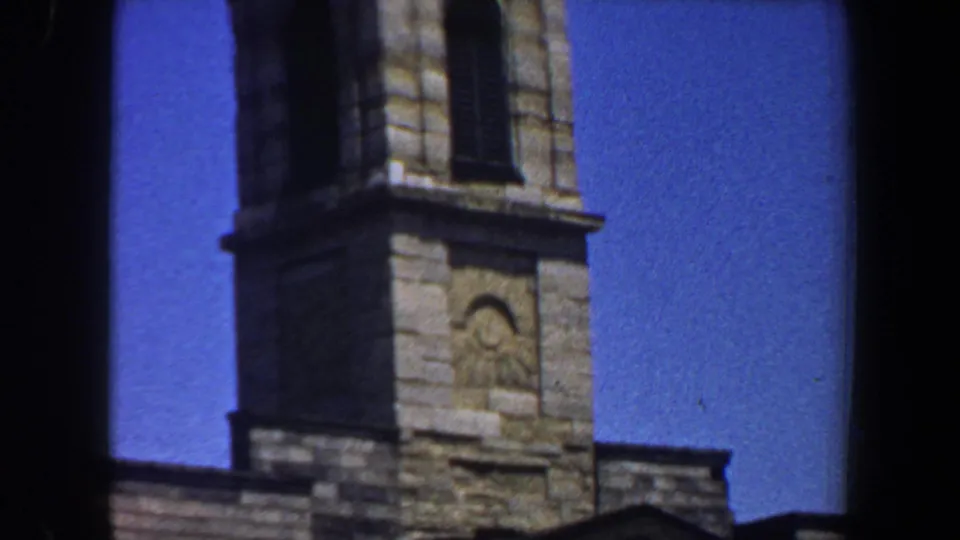 Upper portion of a Saint Louis church tower with stone blocks, arched carving, and blue sky in 1964 home-movie footage.