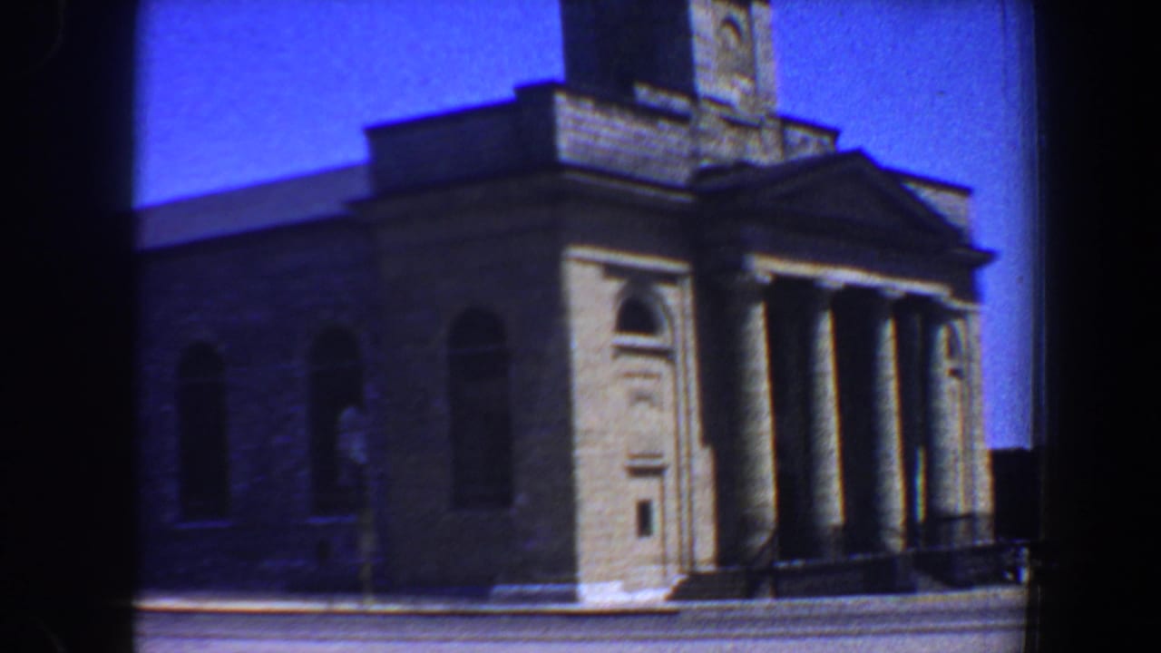 1964 Saint Louis 8mm frame showing a stone building exterior with a columned entrance, arched windows, and bright blue sky.