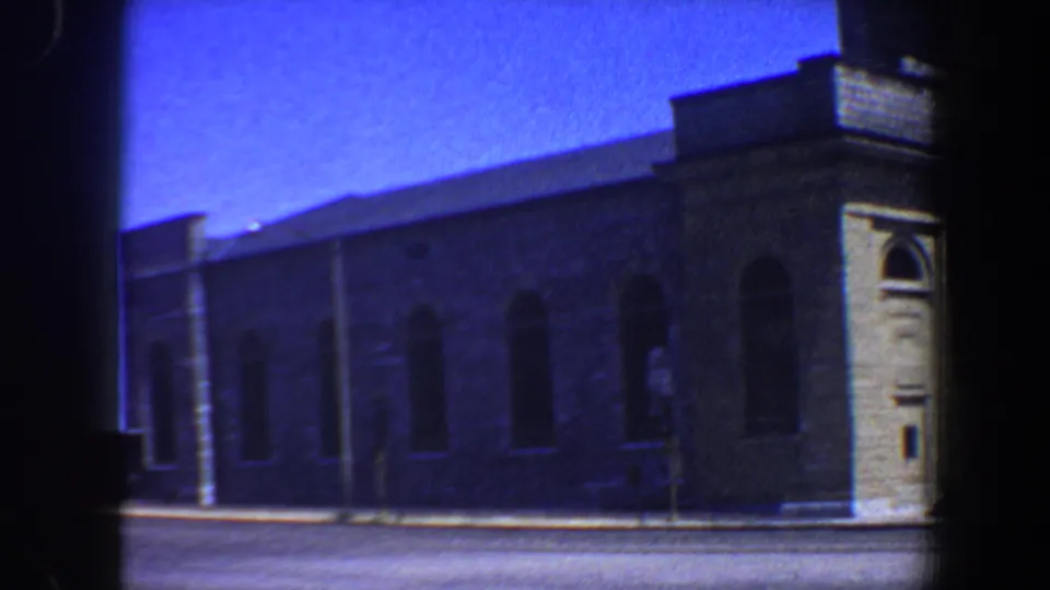 1964 Saint Louis home-movie frame showing the side of a stone building with repeated arched windows and a quiet street in front.