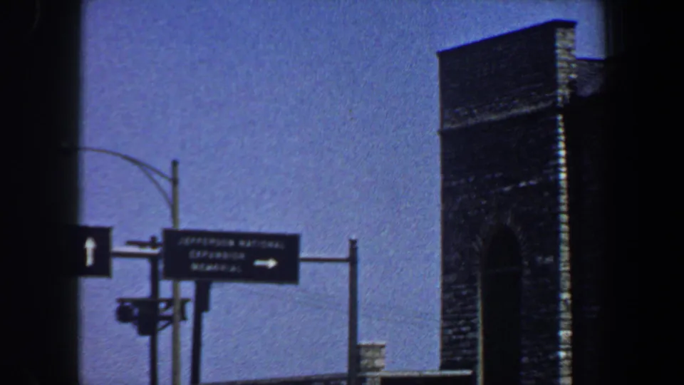 Saint Louis roadway sign for Jefferson National Expansion Memorial near a tall brick or stone structure, filmed in 1964.