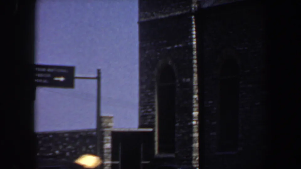 1964 Saint Louis 8mm frame showing a directional sign near a dark brick building with arched architectural openings.