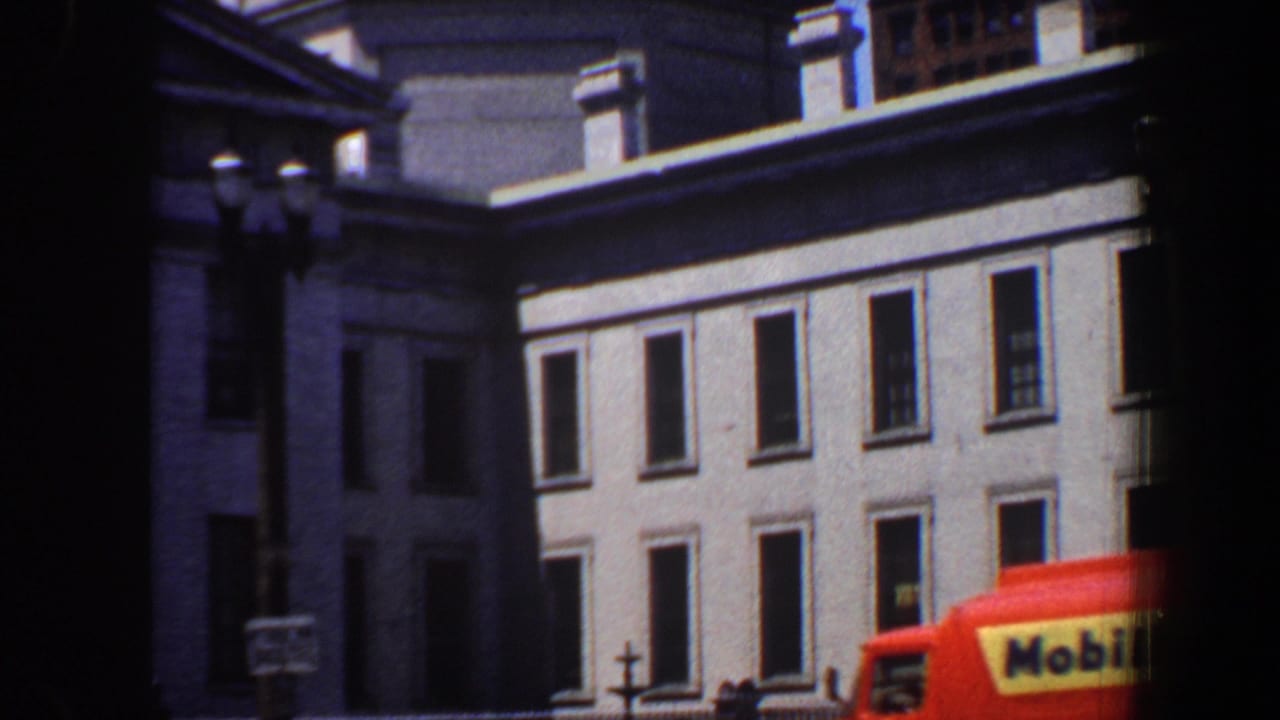 Grey classical building with domed tower and red-orange bus entering the foreground.