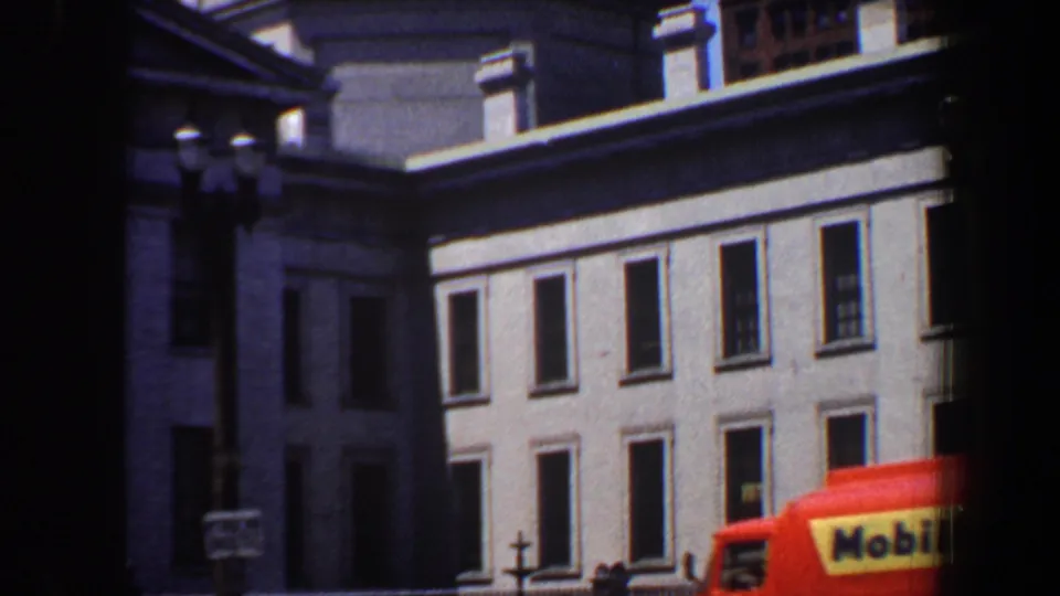1964 Saint Louis street view with a grey classical building, domed tower, lamps, and a red-orange bus partly crossing the foreground.