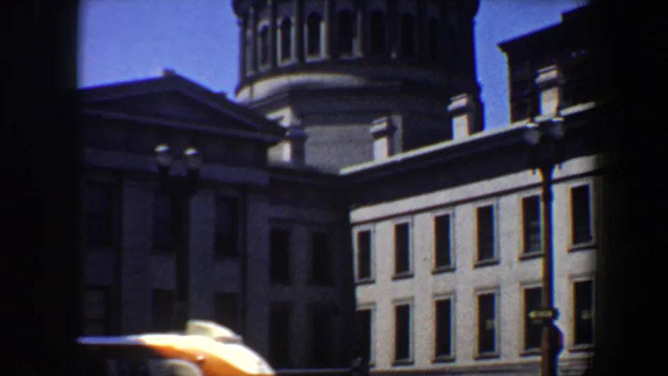Saint Louis street view from 1964 showing a grey stone building and tower while a pale car passes through the foreground.