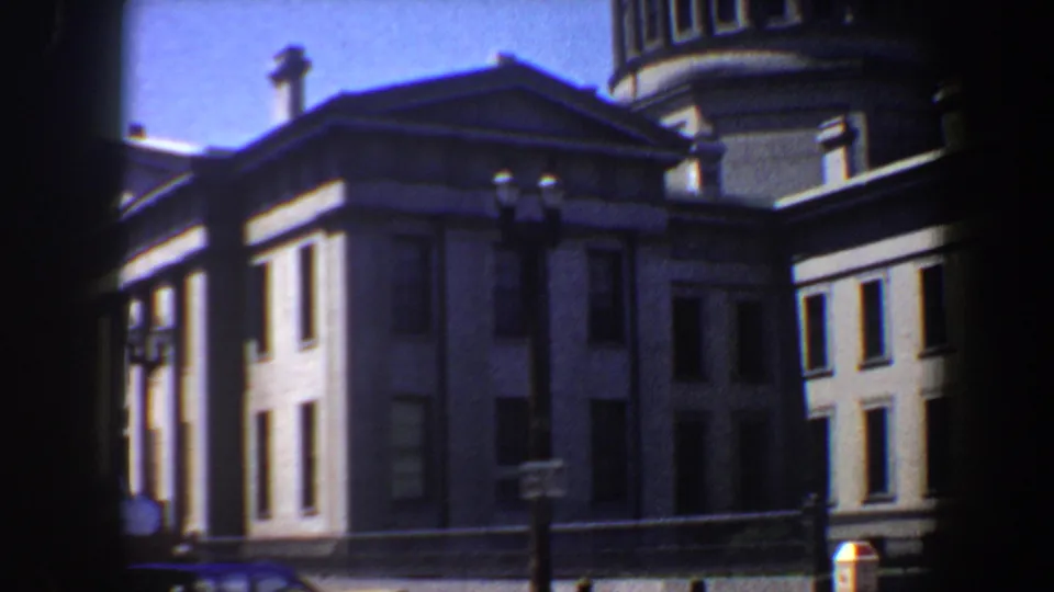 Late frame from the 1964 Saint Louis clip showing the grey building corner, tower, street lamps, and sunlit blue sky.