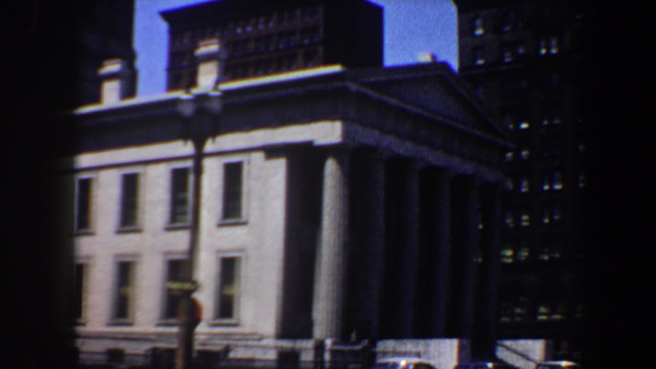 Columned classical building framed by downtown high-rises and cars.