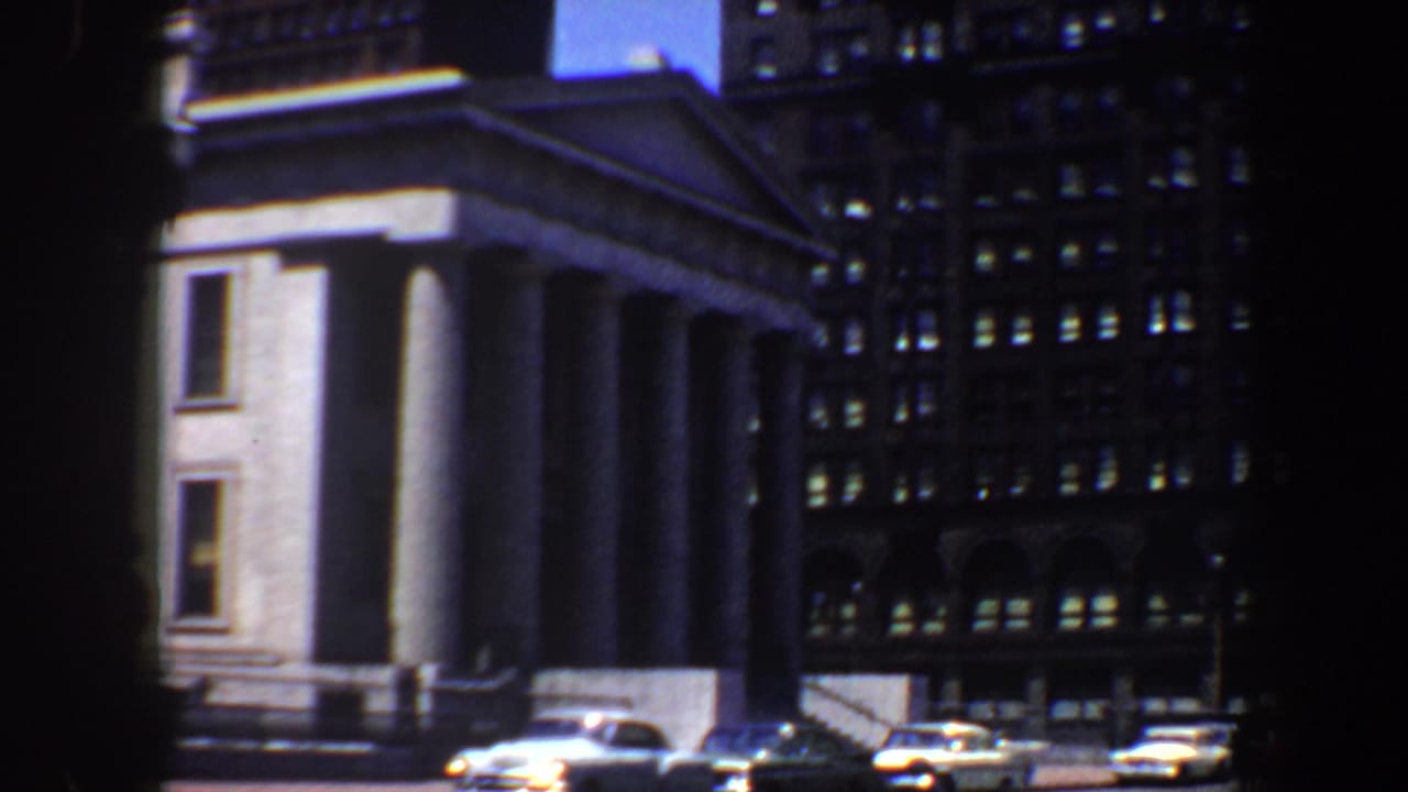 Similar view of the columned building with cars along the street.