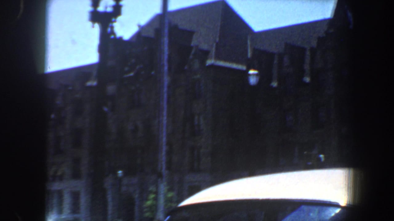 1964 Saint Louis home-movie frame showing a shadowed ornate building, streetlight, blue sky, and white car roof foreground.