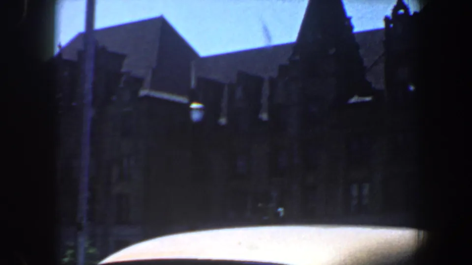 Drive-by 8mm view in Saint Louis, Missouri, with a dark gabled building, pale car roof, and bright blue sky.