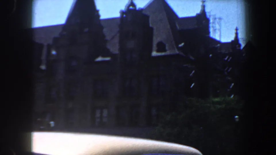 Late frame from the 1964 Saint Louis drive-by, showing a gabled building, white car roof, street pole, foliage, and blue sky.