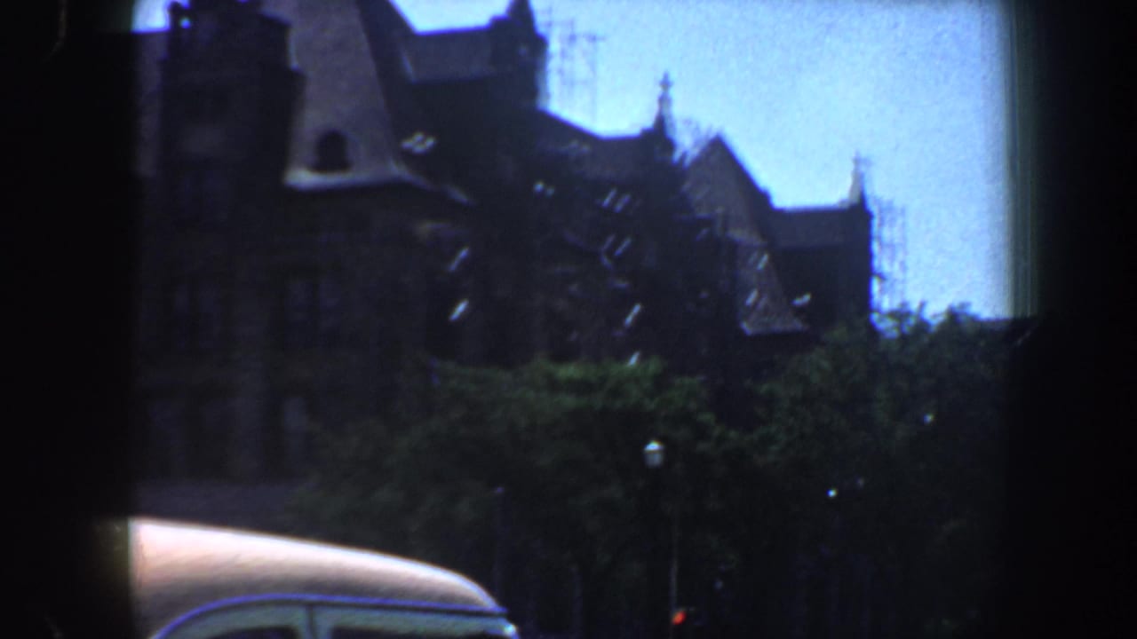 1964 Saint Louis home-movie frame showing a spired stone building behind trees, with the curved roof of a car crossing the lower frame.