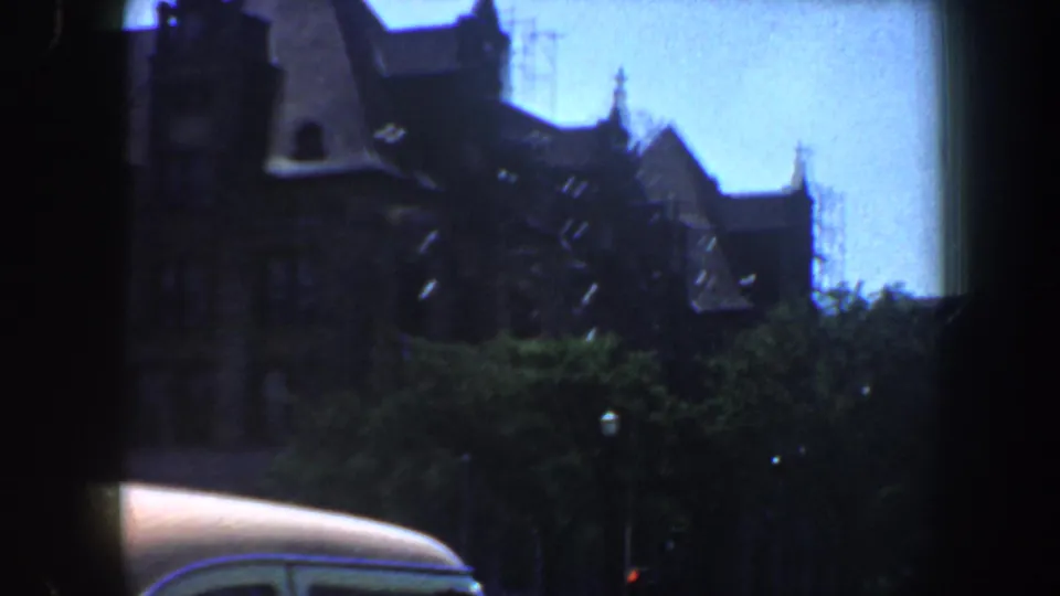 1964 Saint Louis home-movie frame showing a spired stone building behind trees, with the curved roof of a car crossing the lower frame.