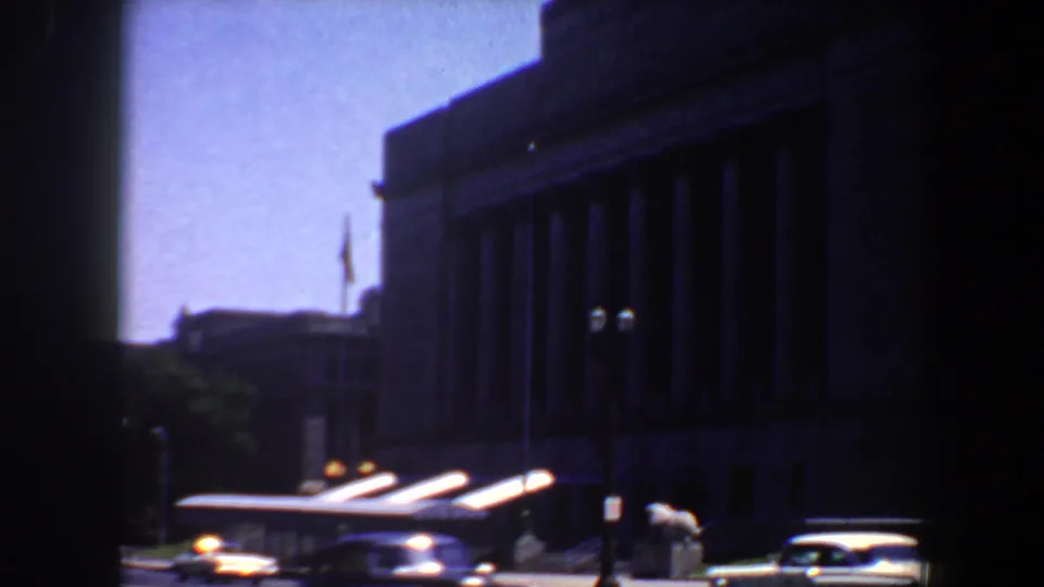 1964 street view of a large Saint Louis building with columns, a white car, and sidewalk activity at a distance.