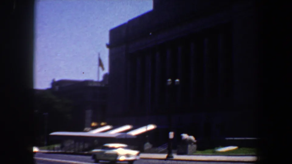 Late frame of the Saint Louis street scene showing the columned facade, flags, streetlamps, and passing cars.