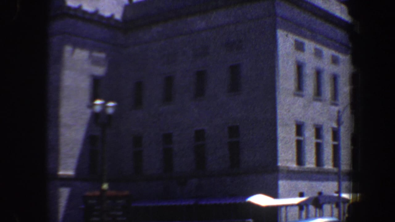 Corner of a large building with window rows, streetlamp, dark sign, and bright awning near the curb.