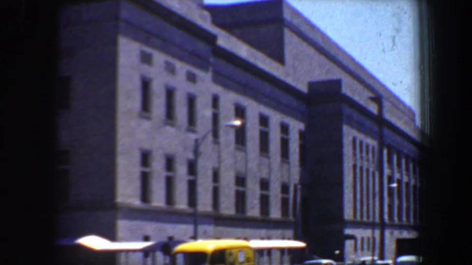 1964 Saint Louis street view of the same large windowed building, with lamps, bright awnings, and a yellow vehicle partly visible below.