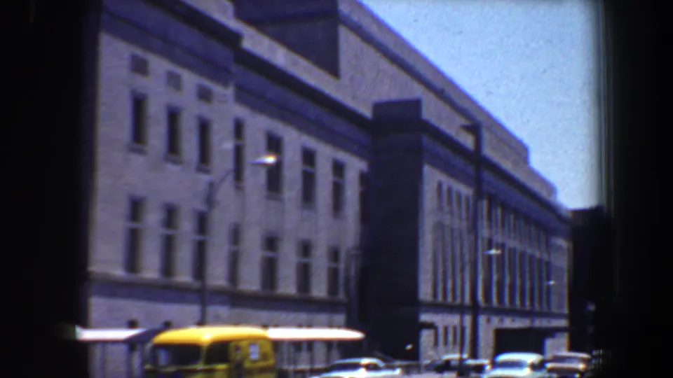1964 Saint Louis street view with classic cars passing a large institutional building and streetlights.