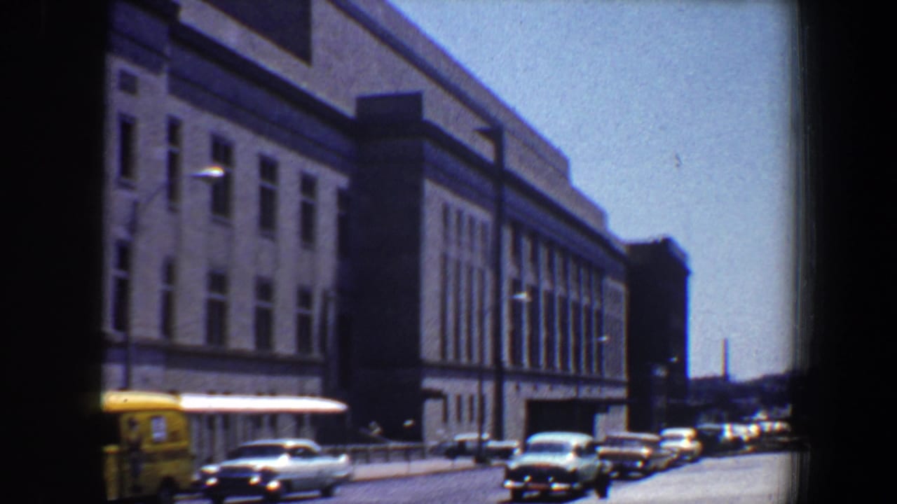 Classic cars and a yellow vehicle pass a large city building.