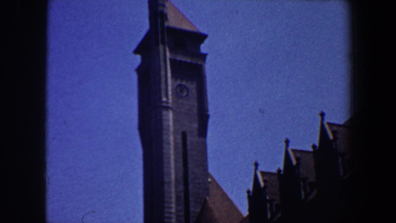 Grey belltower with clock face rising beside dark roof silhouettes against blue sky.