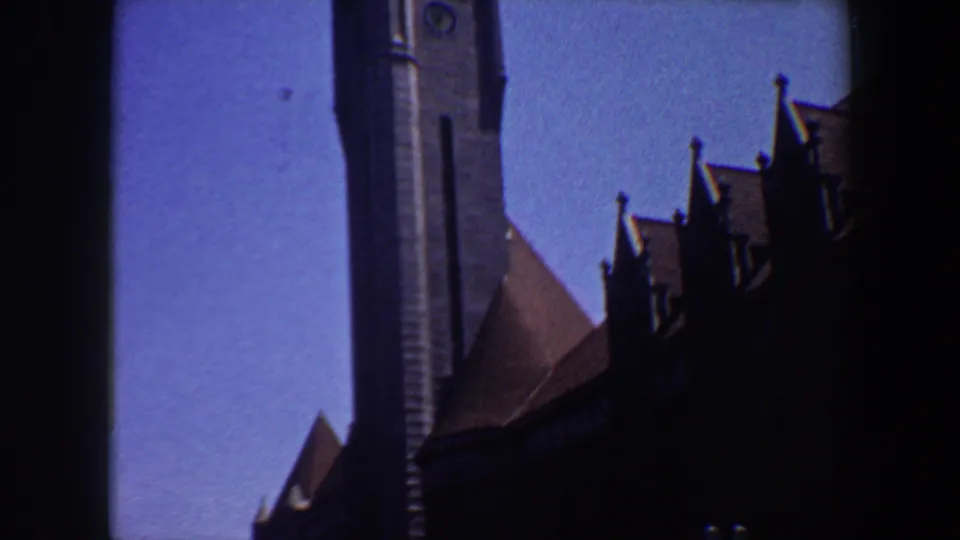 1964 Saint Louis 8mm frame of a grey stone clock tower rising above dark roof silhouettes in bright daylight.