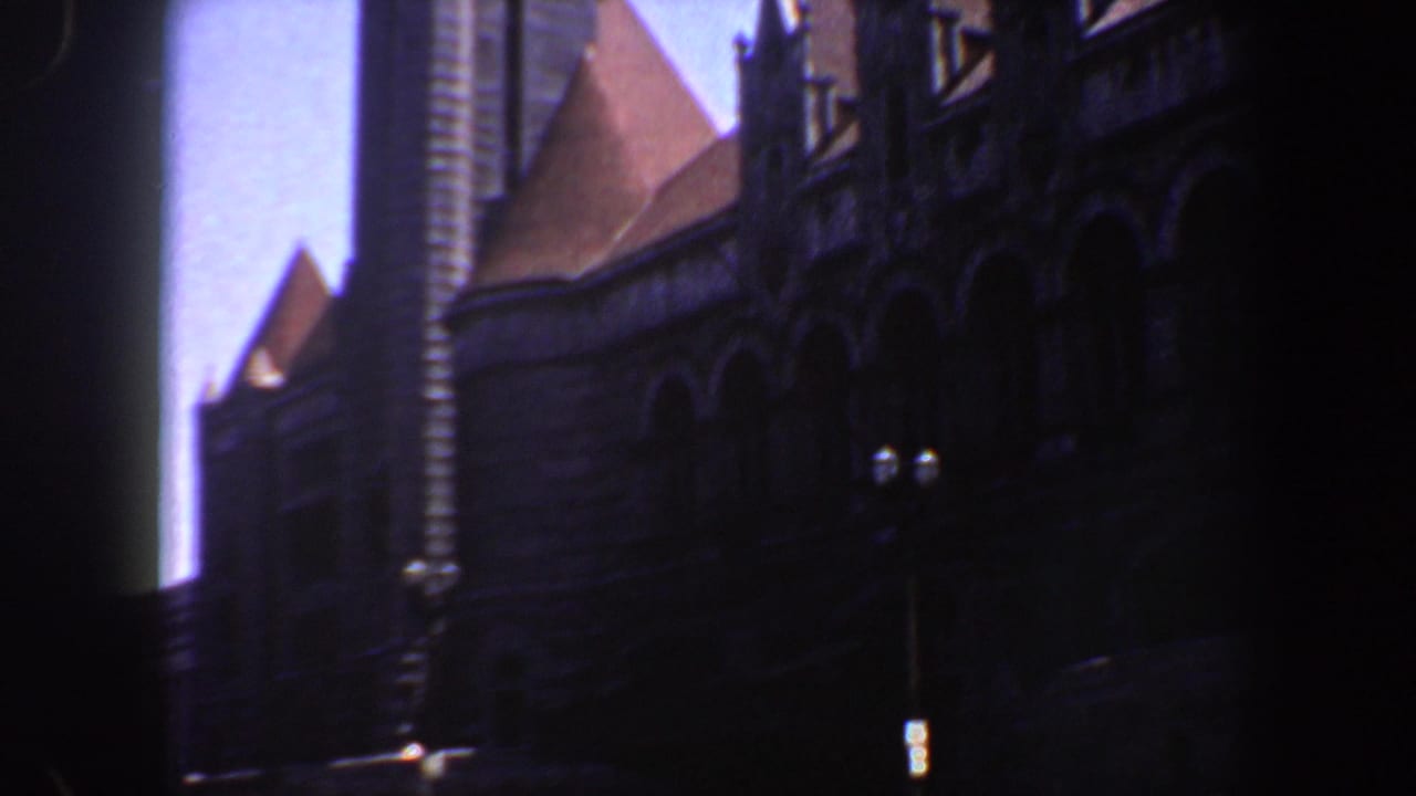 1964 Saint Louis 8mm frame showing a dark stone facade, pointed roof forms, dormers, and a tall vertical architectural element against blue sky.