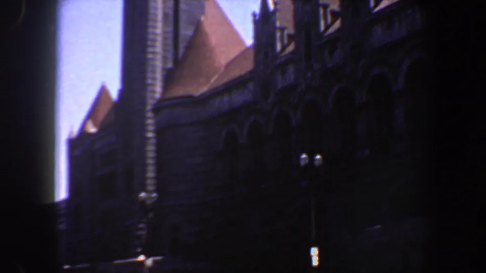 1964 Saint Louis 8mm frame showing a dark stone facade, pointed roof forms, dormers, and a tall vertical architectural element against blue sky.