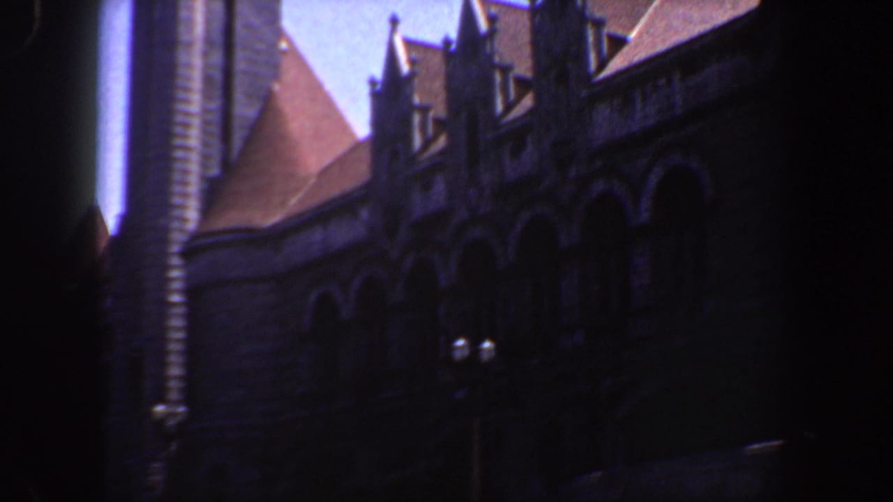 Shadowed building facade with arched stonework, red roof sections, dormers, and a bright sky.
