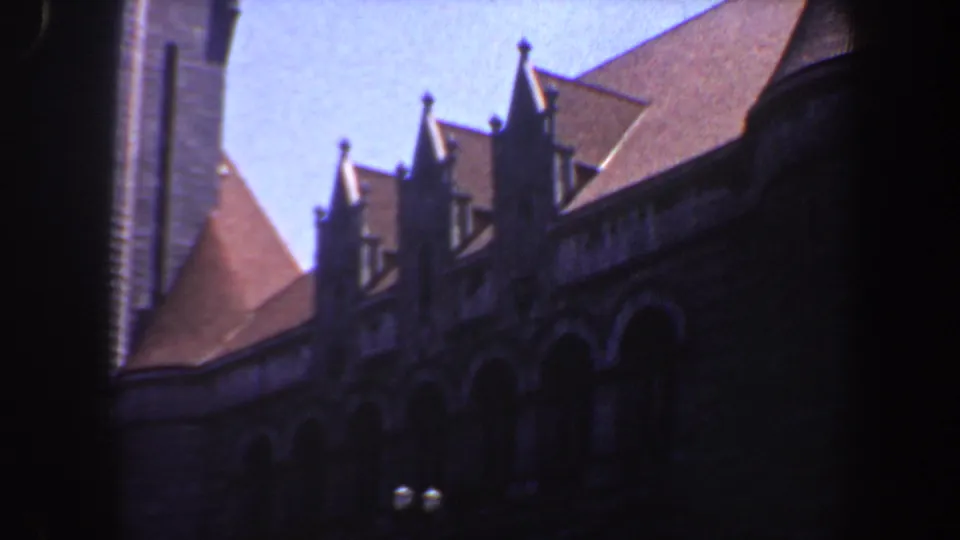 1964 Saint Louis 8mm frame showing a closer view of pointed dormers, steep red roof planes, arched facade details, and a dark foreground edge.