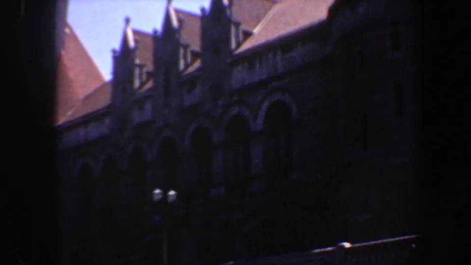 1964 Saint Louis 8mm frame of a shadowed Gothic Revival facade with red roof sections, dormers, arched stonework, and a central dark obstruction.