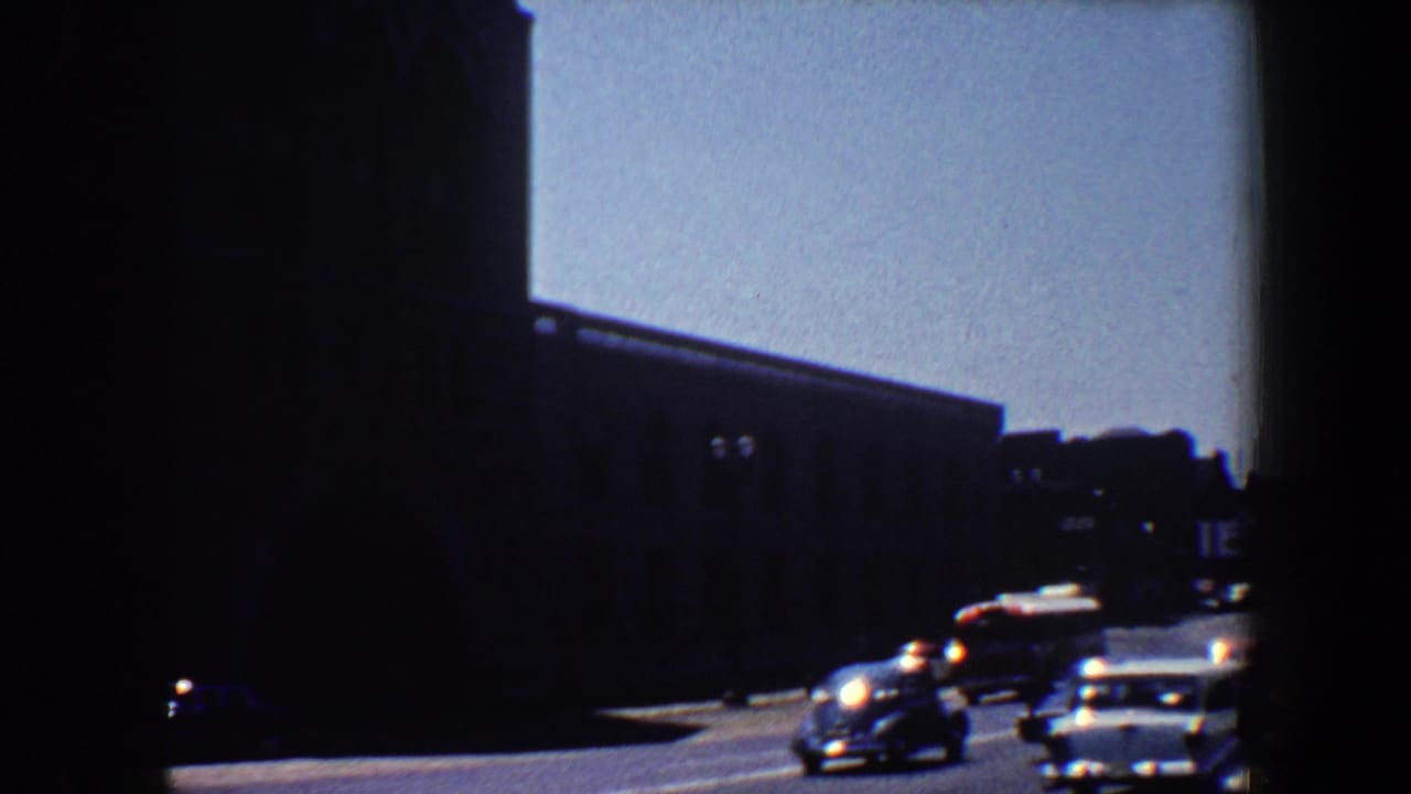 Dark city street with classic cars passing low buildings under bright sky.