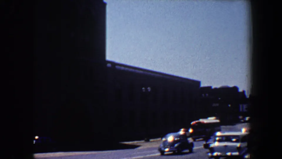1964 Saint Louis 8mm frame showing traffic moving past shadowed urban buildings on a sunny street.