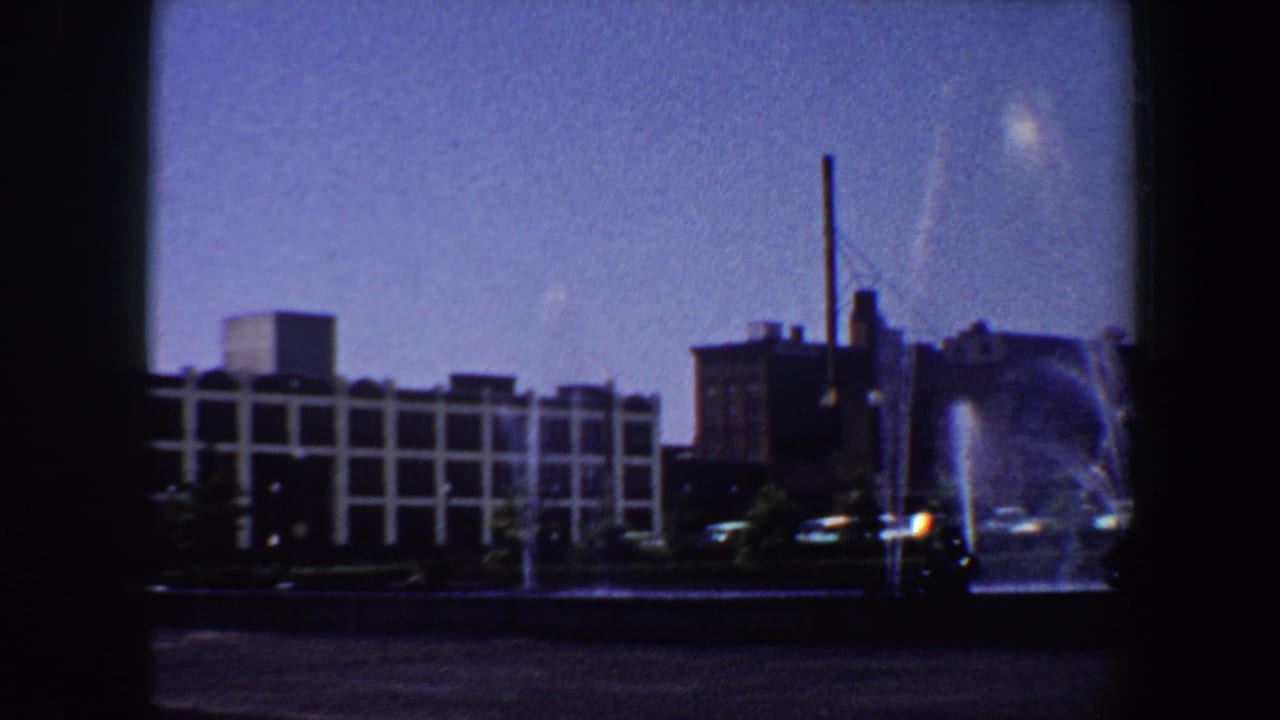 1964 Saint Louis view of a public fountain with mid-century buildings and a tall smokestack beyond it.