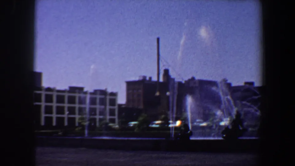 1964 Saint Louis frame showing a fountain, white grid-patterned building, smokestack, and bright sky from a handheld view.