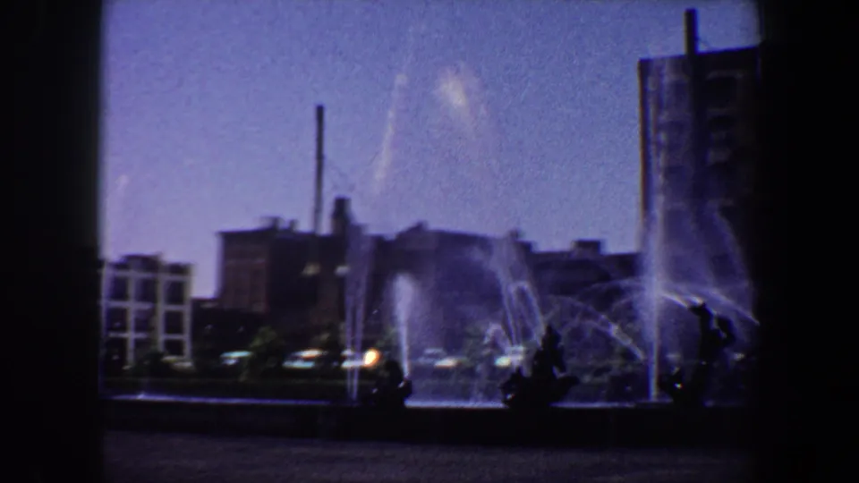 1964 Saint Louis 8mm frame of a civic fountain, streetlamp, smokestack, and surrounding downtown buildings.
