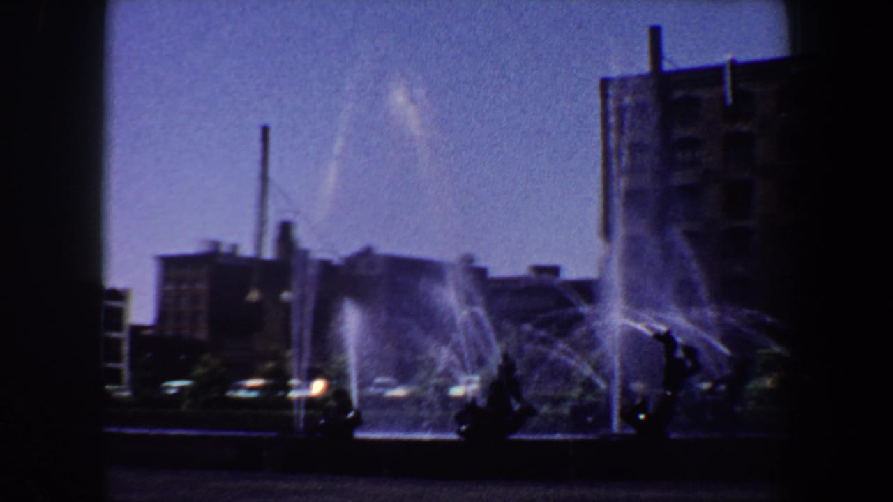 City fountain spraying in a plaza with dark sculptures, cars, and brick buildings behind it.