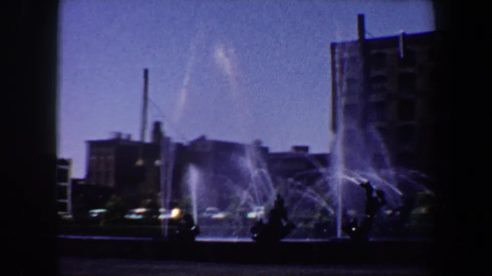 1964 Saint Louis home-movie frame showing a public fountain, silhouetted sculpture forms, cars, brick buildings, and a tall vertical stack.