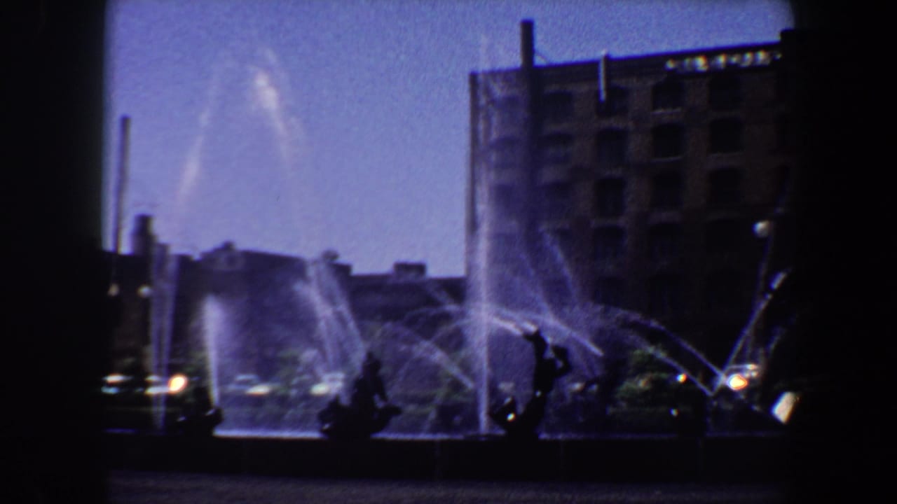 Wide view of the fountain plaza with water jets, sculpture silhouettes, cars, and a brick building.