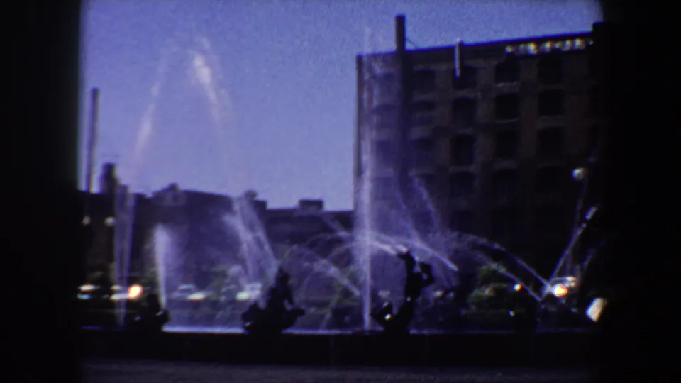 1964 Saint Louis frame with multiple fountain jets crossing the foreground before dark sculptures and a brick urban building.