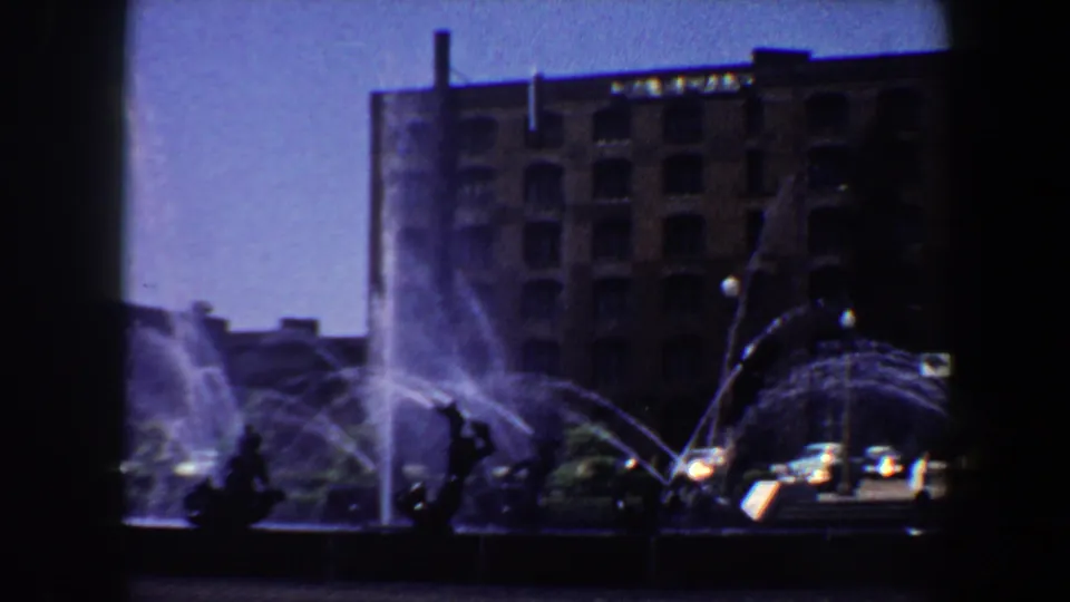 Saint Louis 1964 fountain scene with strong spray, shadowed sculpture shapes, city buildings, and a vehicle near the plaza edge.
