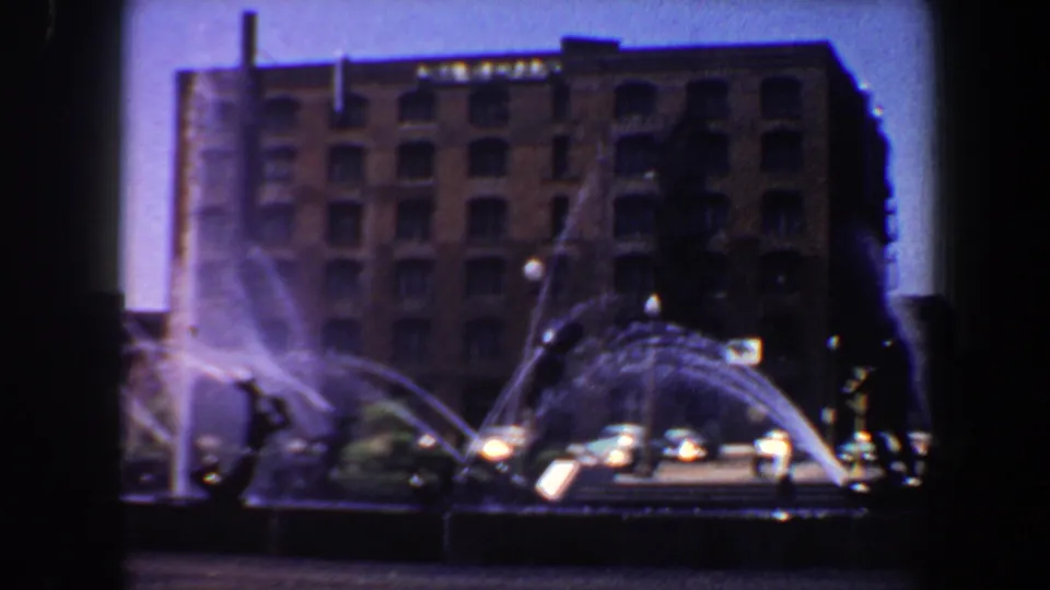 Late frame of the 1964 Saint Louis fountain plaza, with bright water arcs, sculpture silhouettes, cars, and a brick downtown facade.