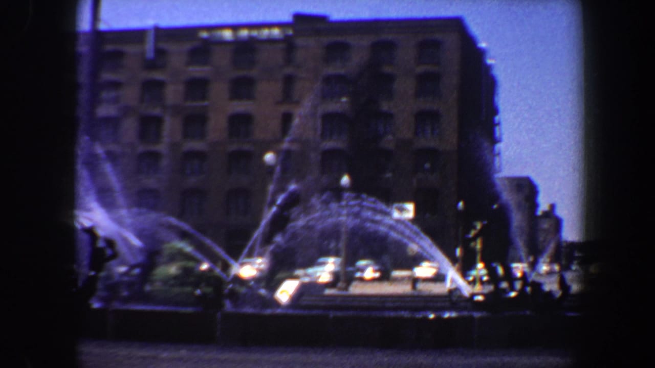Street fountain spraying in front of a brick multi-story building with cars and pedestrians nearby.