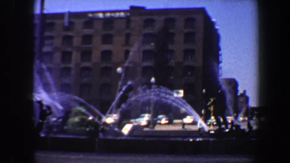 1964 Saint Louis street view with arcing fountain water, a brick high-rise backdrop, passing cars, and small pedestrian silhouettes.