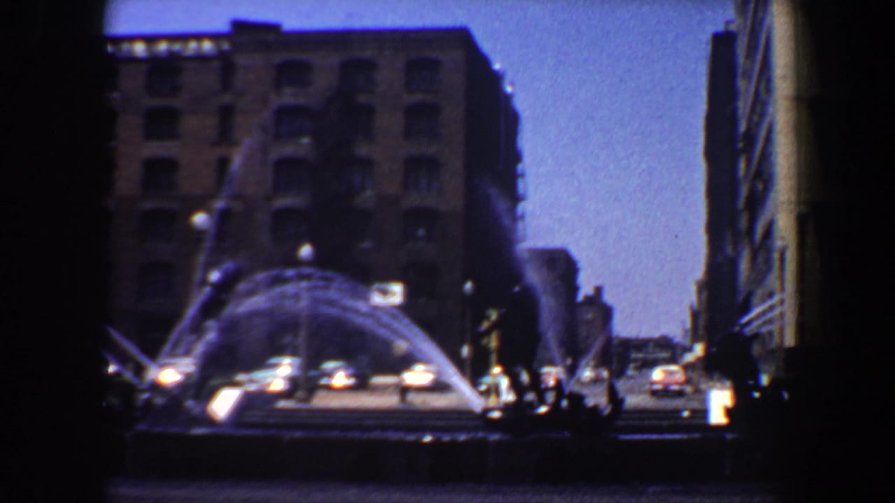 Large fountain sprays across the foreground while cars pass beside urban buildings.