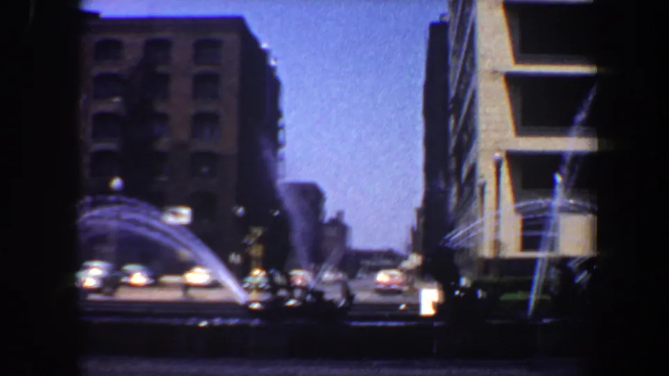 1964 Saint Louis street fountain framed by a brick high-rise, a modern adjacent building, passing cars, and small figures near the plaza.