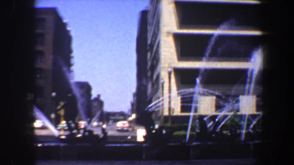 1964 Saint Louis view of fountain jets near a modern building facade, with traffic and silhouetted pedestrians along the street.