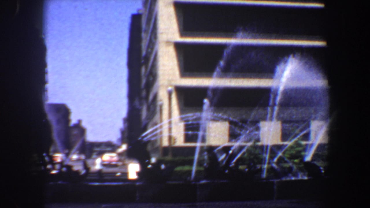 Fountain jets arc in front of a modern building and street traffic.