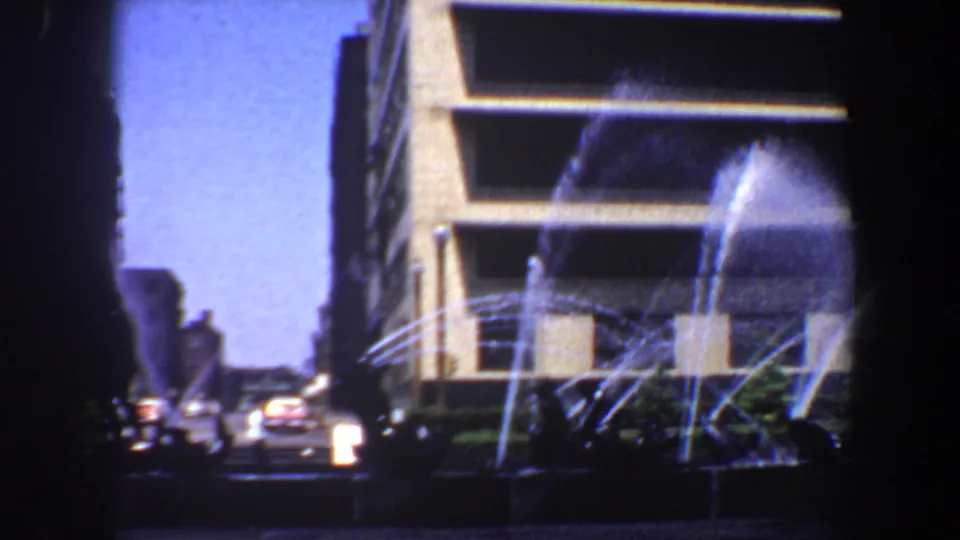 1964 Saint Louis 8mm frame showing a public fountain spraying before a modern building with cars along the street.