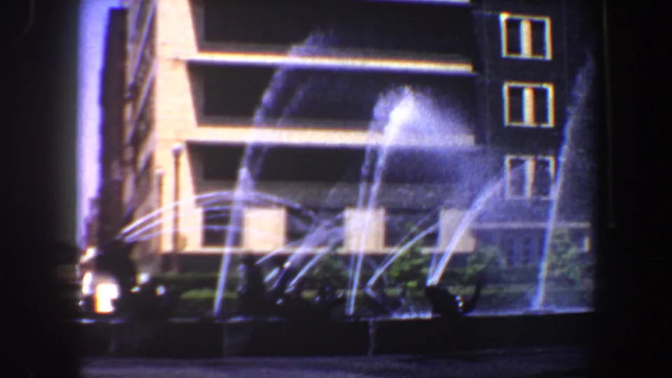 1964 Saint Louis frame with multiple fountain streams, shrubs, a low basin, and a tiered modern building exterior.