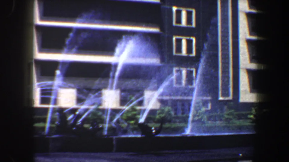 1964 Saint Louis 8mm frame showing bright fountain jets in front of a modern building, landscaping, and cars.